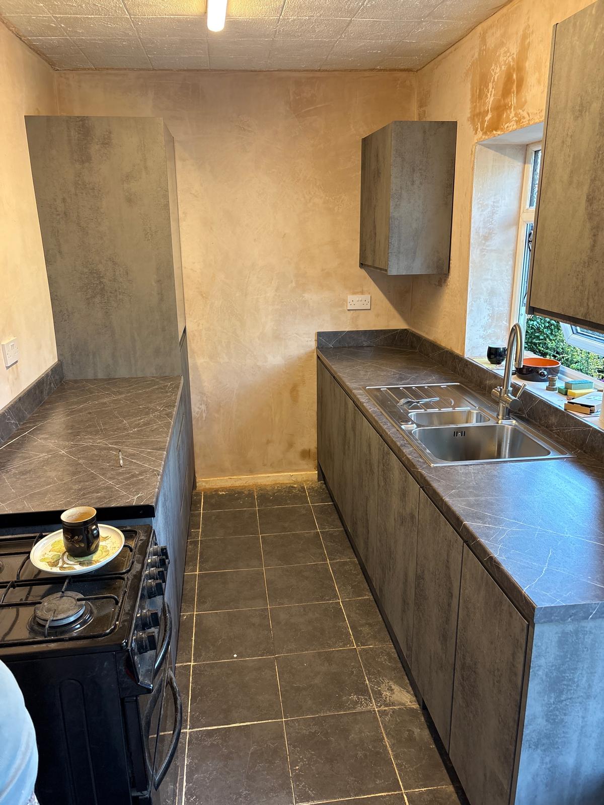 Complete strip-out of a tired galley kitchen — old units, tiling and finishes removed, walls re-plastered, new floor tiles laid and contemporary stone-effect units installed with a new sink, tap and worktops.