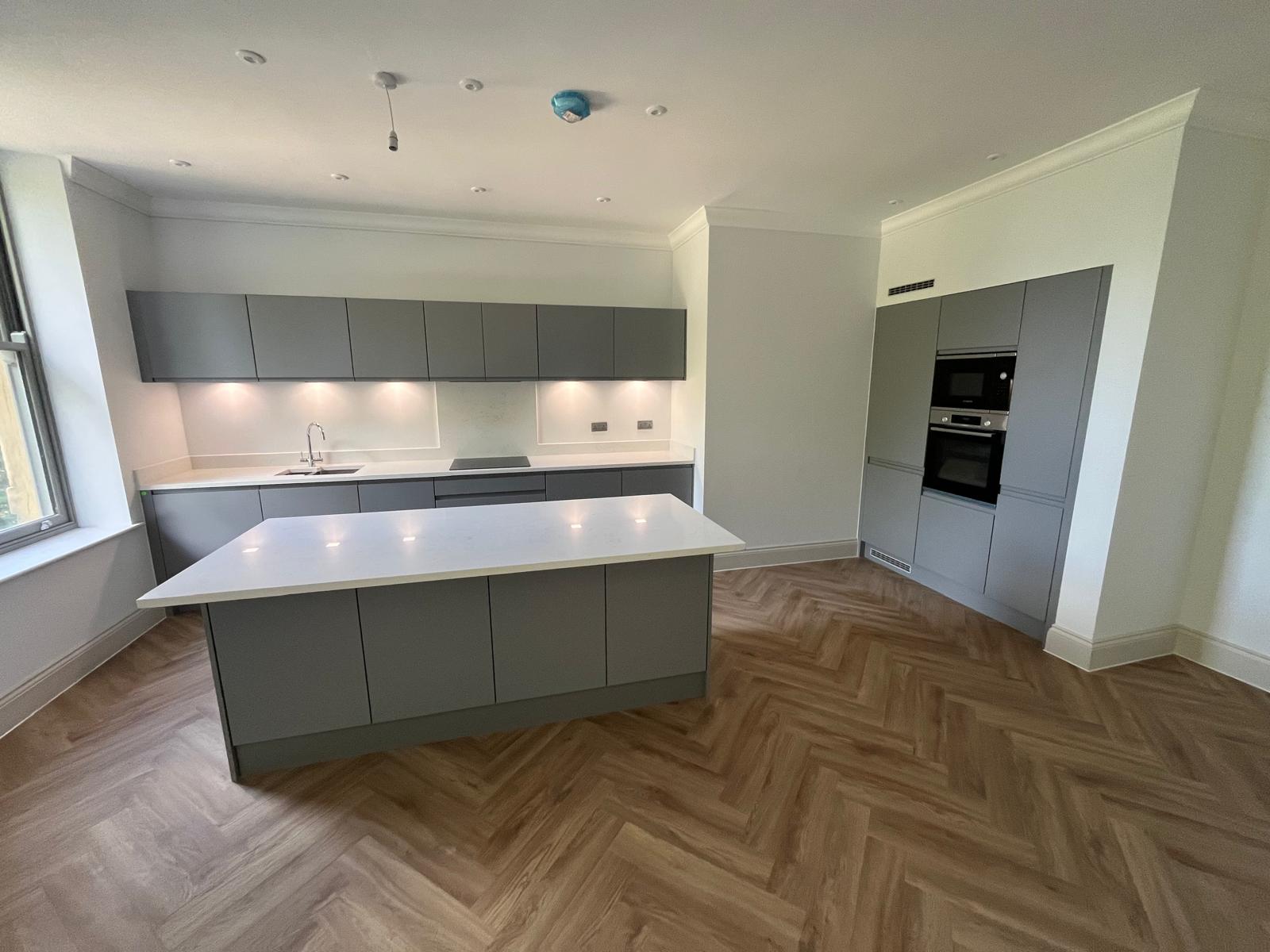 Sleek handleless grey kitchen with white quartz worktops, integrated appliance tower and herringbone LVT flooring throughout.