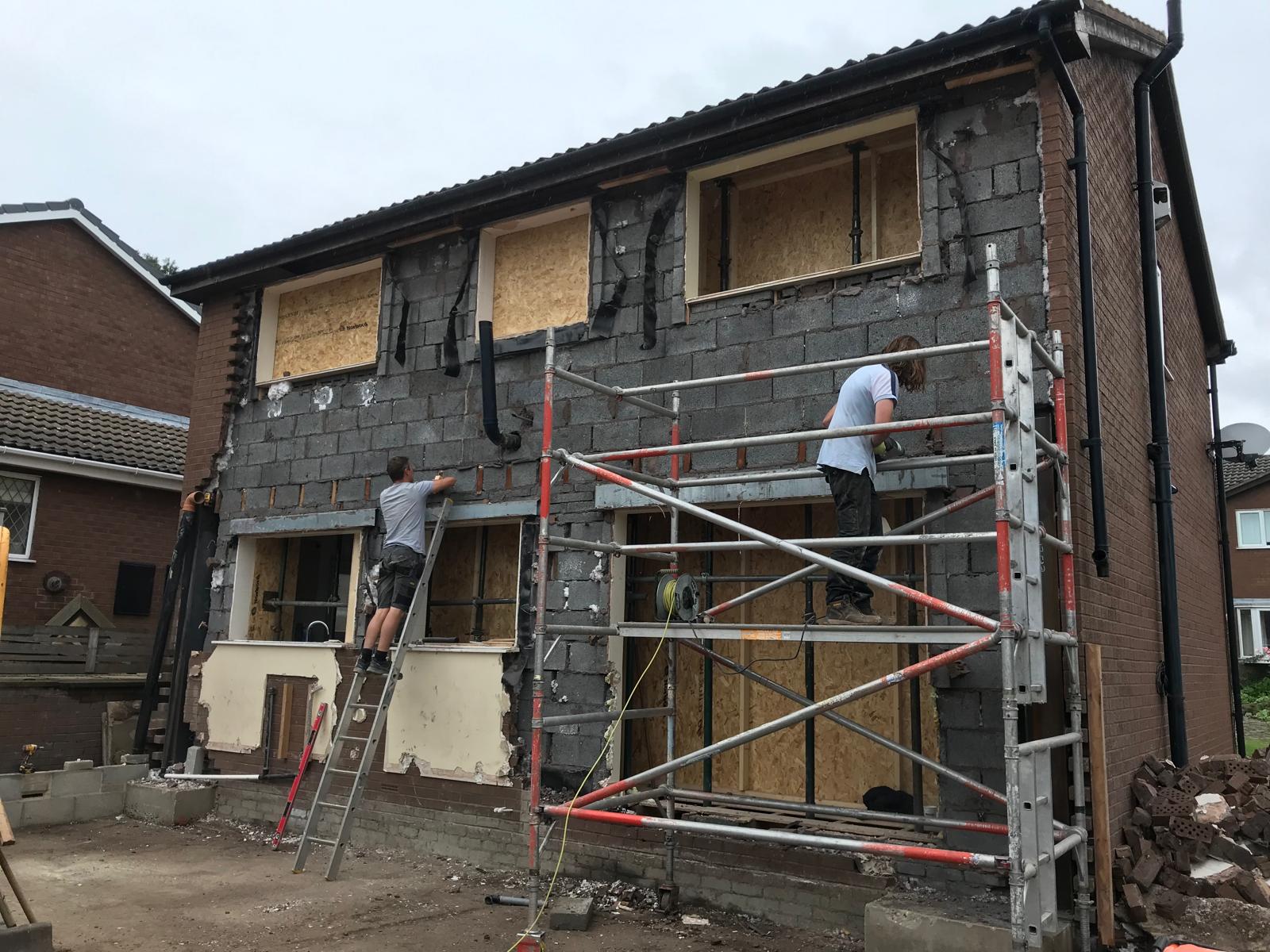 Bricklayers cutting in new window openings and lintels on a two-storey side extension, with scaffold towers either side and original render being trimmed back.